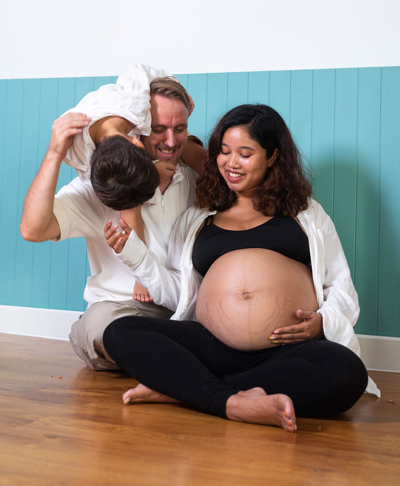 A pregnant mother, a father, and a young son hug in an indoor environment.